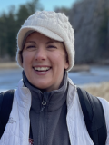 A photo of Kateri McRae, a woman smiling at the camera while wearing a white coat and a cream-colored knit hat